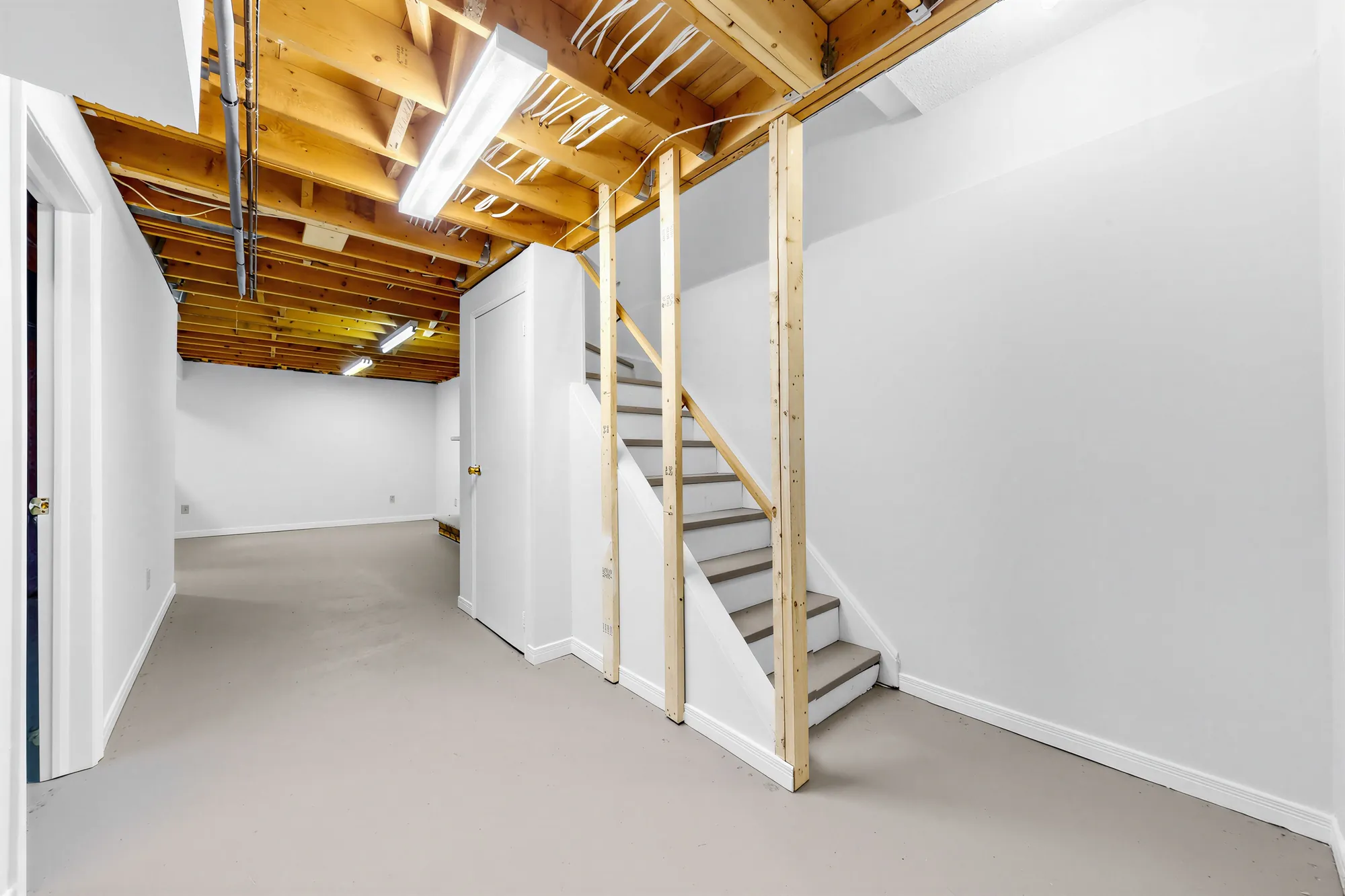 The foot of the basement stair: open joists, an unfinished stud wall, a painted floor, a door at the left