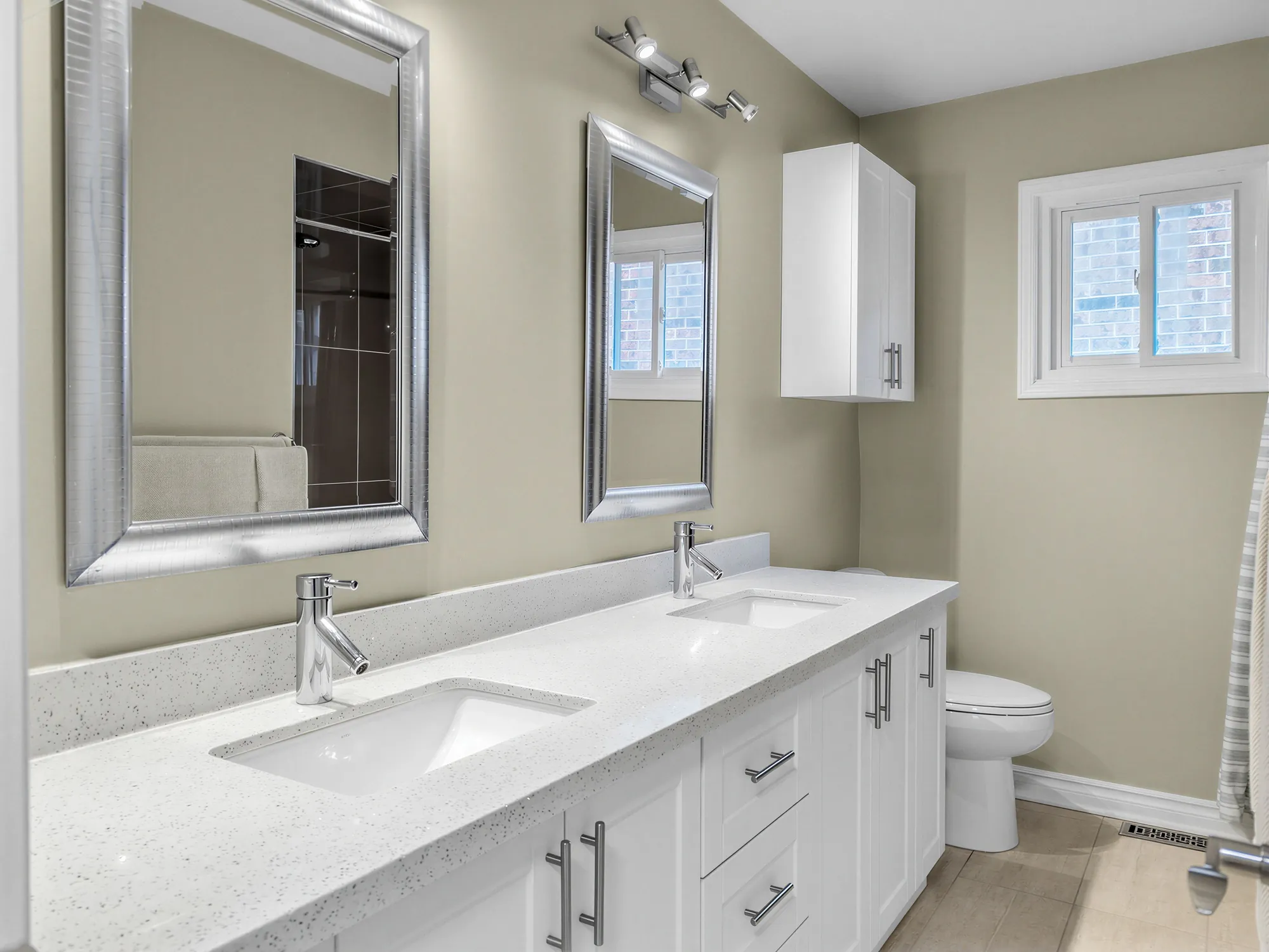 The double vanity in white with a quartz top, two framed mirrors, a toilet, a window, olive walls
