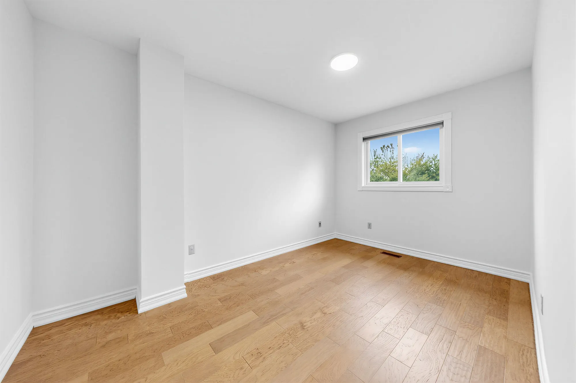 The same bedroom toward its window, a wall jog at the left, hardwood floors