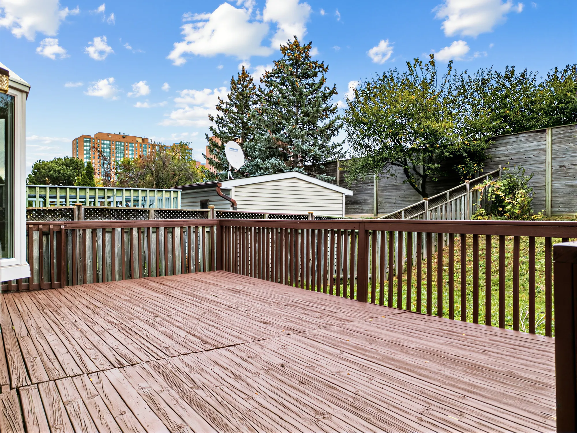The deck at the back: wide boards and a wood railing, the fenced yard, a neighbour&rsquo;s shed, trees and a tower beyond