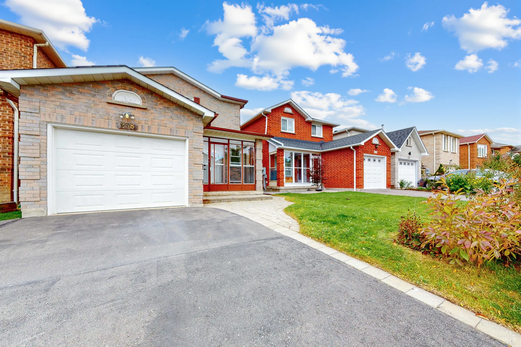 The house from the street at an angle: the garage with its white door, the paved drive, an interlocking walk, the lawn and the enclosed porch, neighbouring houses along the street, a blue sky with clouds