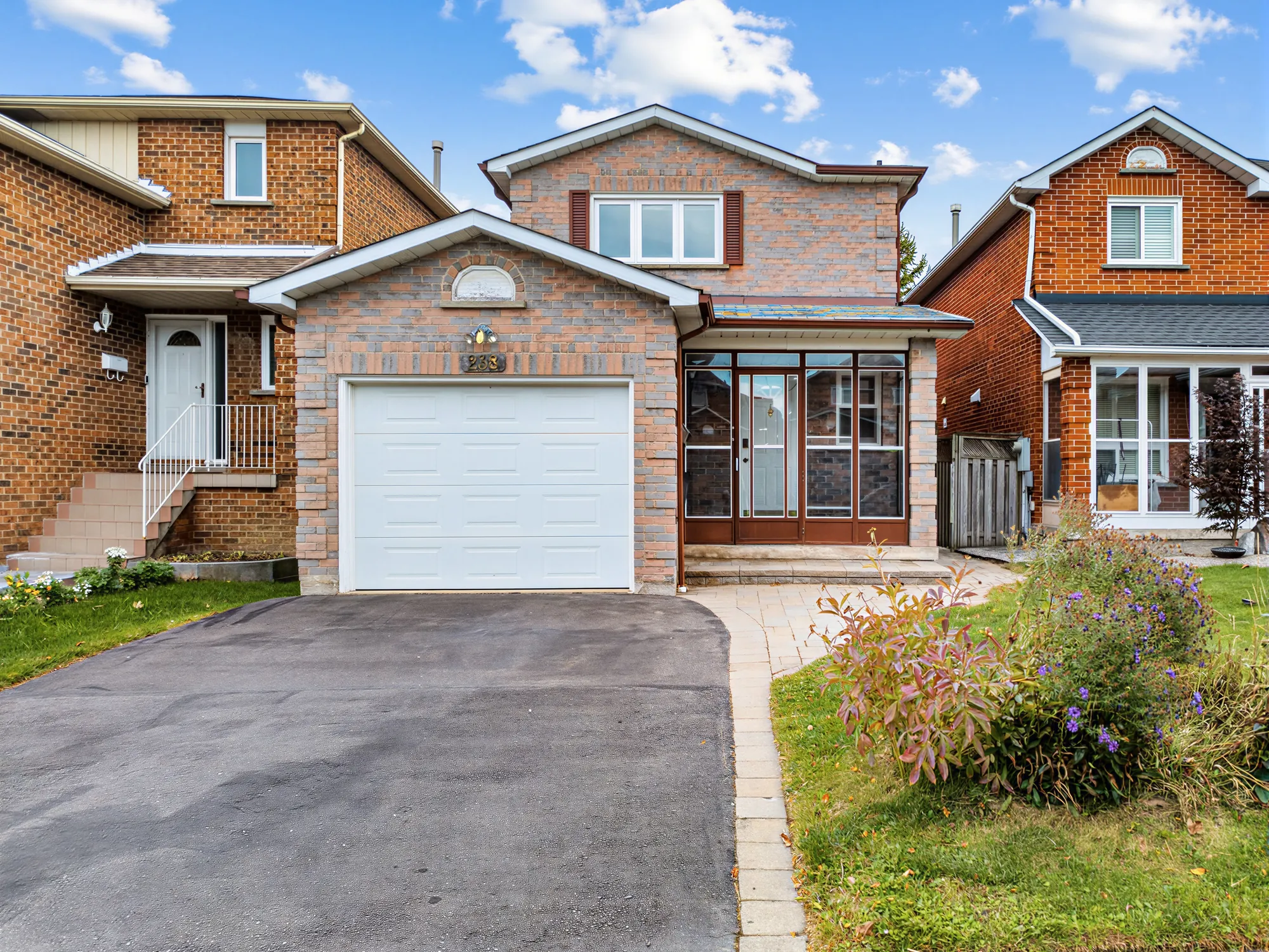 The front of the house by day: two storeys of grey and rose brick, a single garage with a white door under a gable with an arched window and the house number, an enclosed porch glazed on the front at the right, a paved drive and a bed of asters