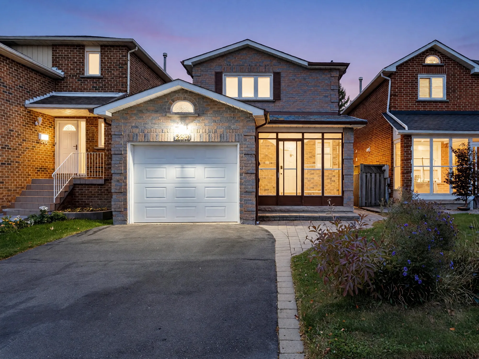 The front of 238 Lech Walesa Drive at dusk, an edited photograph: two storeys of brick, a single garage with a white door, an enclosed porch lit at the right, a paved drive