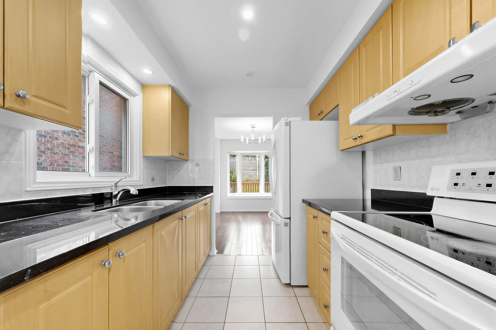 The kitchen: maple cabinets on both sides, black granite counters, a double sink under a window, a white range under a hood, a white fridge, the dining room window at the far end