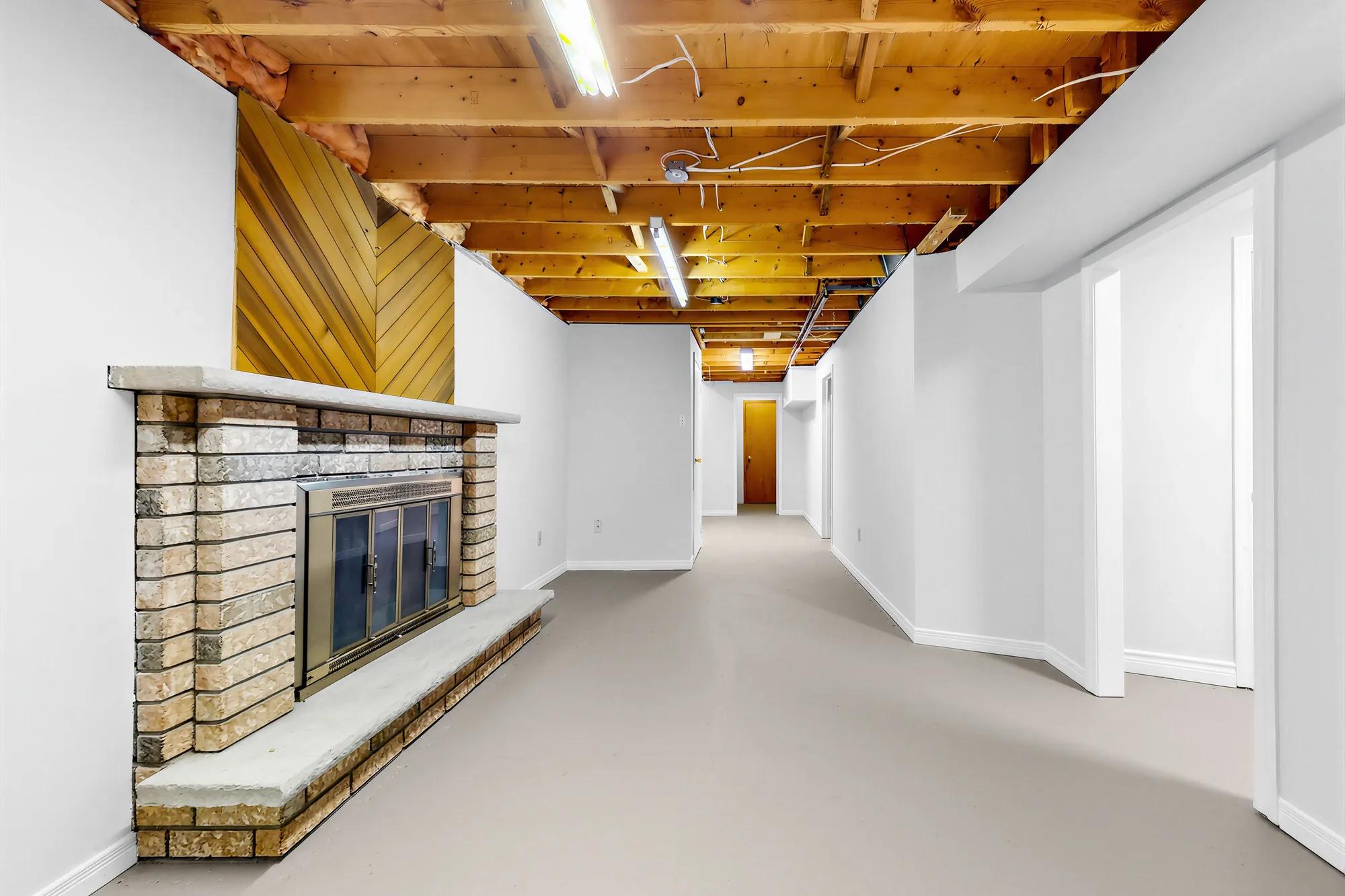The recreation room in the basement: a brick fireplace with a raised hearth under a wood-panelled chimney breast, a painted floor, the ceiling joists open, the hall beyond