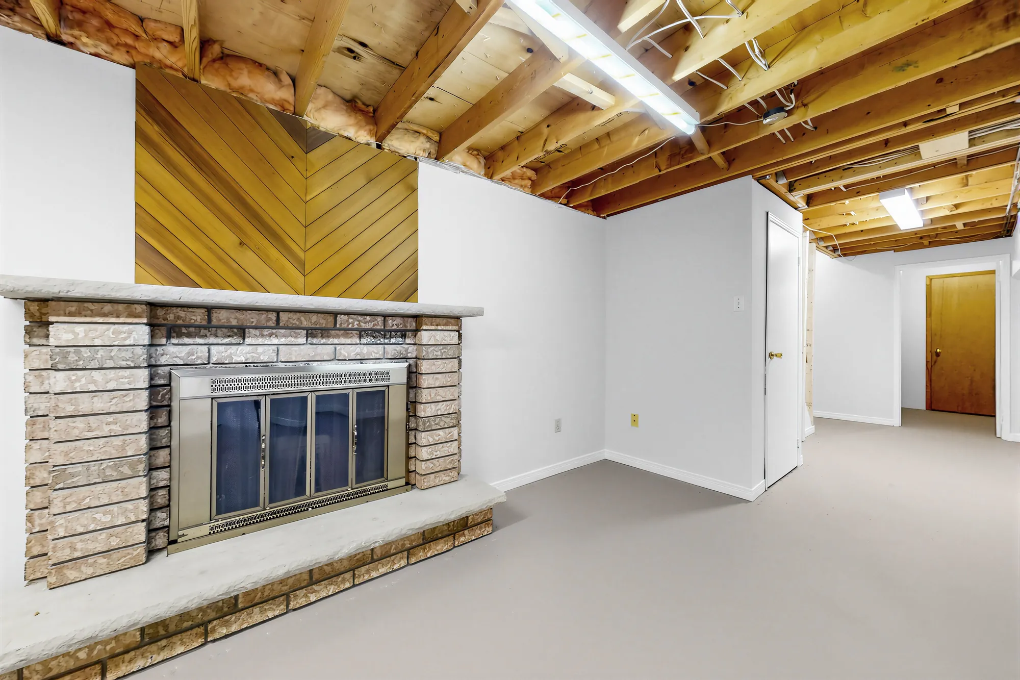 The fireplace from the side, the wood panelling above it, the open ceiling and fluorescent lights, a door at the far end